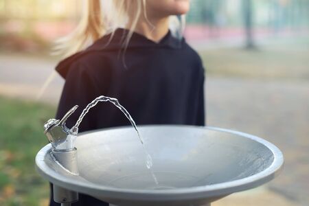 Cute adorable caucasian blond little thirsty school girl drinking water from public potable fountain faucet in city park on bright hot summer day outdoors. Children dehydration at heat seasonの写真素材