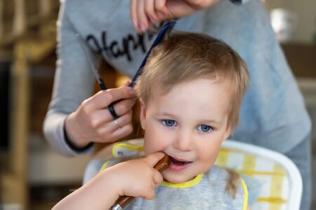 Close-up young adult caucasian mother making haircut fot cute adotrable son toddler boy at home due quarantine and lockdown. Mom cutting hair of child with scissors and hairbrushの写真素材