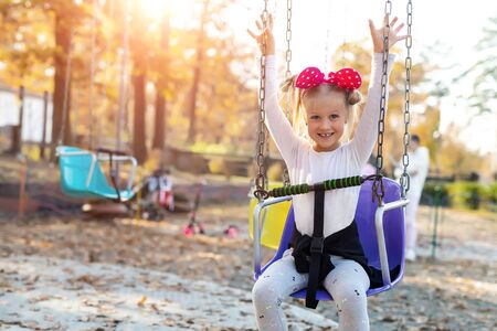 Little cute adorable caucasian blond school girl enjoy having fun sitting and riding old chain swing carousel at city theme amusement park. Happy child portrait outdoors at sunset evening sun lightの写真素材