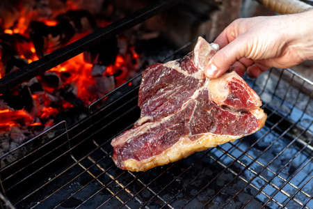 Close-up man hand putting big tasty juice raw t-bone beaf steak on grill at barbecue with bright hot burning charcoal at brazier. BBQ meal cooking background.の写真素材