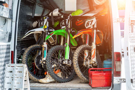 Three bright dirty sport motocross motorcycles stand in tail of technical support assistance van in team camp during extreme offroad championship. Mobile cycle service, maintenance transport concept.の写真素材
