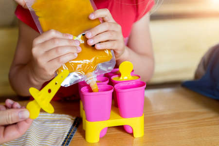 Cute adorable young caucasian little girl kid sitting at table home yard and have fun making with mom homemade fruit puree icecream. Young happy child enjoy cooking with mother outside. Family loveの写真素材