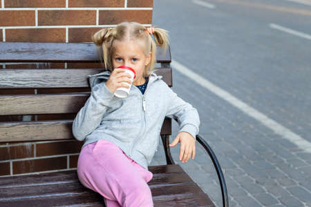Cute adorable little caucasian blond kid girl enjoy sitting om wooden bench and drink hot chocolate drink with paper cup. Beatiful happy smiling 7 years old child portraitの写真素材