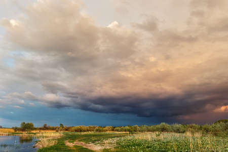 Dramatic dark cloudy thunder sky over dry grass field at pond shore. Rural countryside rainy ominous...