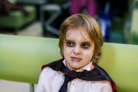 Cute adorable caucasian kid boy in dracula vampire costume and face painting makeup sitting indoors during children halloween party celebrationの写真素材