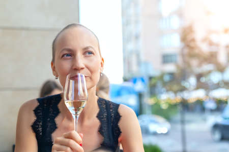 Young adult beautiful attractive stylish short haired woman enjoy sitting in street urban cafe, drinking white wine looking with hope. Positive deep in thought girl portrait with champagne glassの写真素材