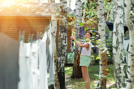 Candid real life portrait of young adult beautiful attractive caucasian woman hanging up fresh washed family clothes on birch tree clothesline with pins at home yard on bright sunny day outdoorsの写真素材