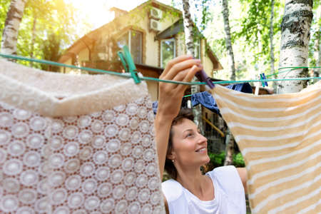 Candid real life portrait of young adult beautiful attractive caucasian woman hanging up fresh washed family clothes on birch tree clothesline with pins at home yard on bright sunny day outdoorsの写真素材
