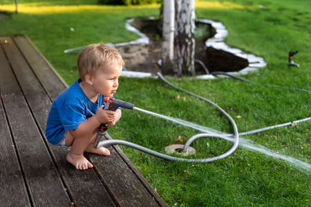 Cute adorable caucasian blond toddler boy enjoy having fun watering garden flower and lawn with hosepipe sprinkler at home backyard at sunny day. Child little helper learn gardening at summer outdoorの写真素材