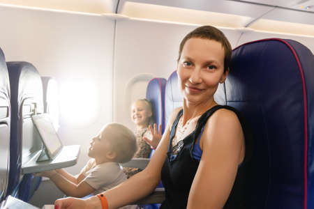 Young adult beautiful attractive caucasian mother traveling with cute little adorable sibling boy and girl by plane. Mom daughter and son sit inside aircraft during flight. Happy family trip conceptの写真素材