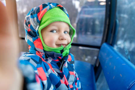 Cute adorable happy caucasian smiling small toddler kid boy enjoy ascent sitting inside ski lift gondola cable car and making selfie camera shot by smartphone. Winter travel with children concept.の写真素材