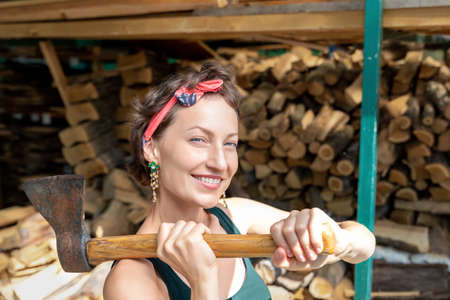 Young adult beautiful happy attractive caucasian brunette woman portrait have fun enjoy holding old rusty ax chopping firewood at wood store on backyard. Female person vintage retro pinup style.の写真素材