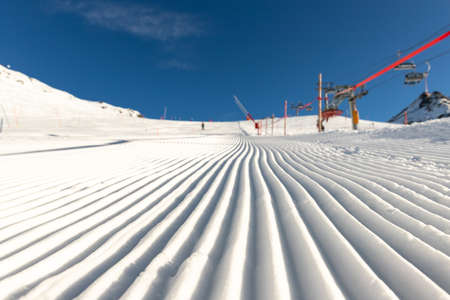 Close-up straight line rows of freshly prepared groomed ski slope piste with bright shining sun and clear blue sky background. Snowcapped mountain downhill landscape at europe winter skiing resort.の写真素材