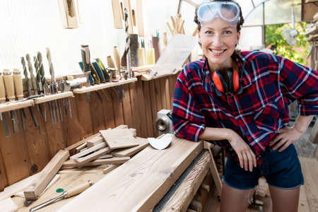 Young beautiful handy professional happy female strong carpenter portrait wearing protective goggles working in carpentry diy workshop against wood. Confident engineer. Women male hobby at workbench.の写真素材
