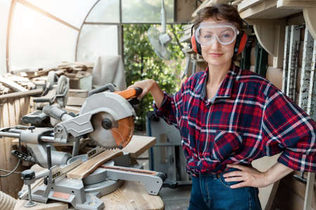 Young beautiful handy professional happy female strong carpenter portrait wearing protective goggles working in carpentry diy workshop with circular saw. Confident Women male hobby at workbench.の写真素材