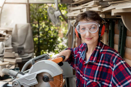 Young beautiful handy professional happy female strong carpenter portrait wearing protective goggles working in carpentry diy workshop with circular saw. Confident Women male hobby at workbench.の写真素材