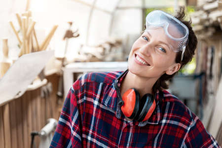 Young beautiful handy professional happy female strong carpenter portrait wearing protective goggles working in carpentry diy workshop against wood. Confident engineer. Women male hobby at workbench.の写真素材