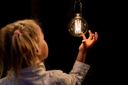 Cute adorable caucasian blond girl portrait smiling and holding in hand one of hanged edison light...