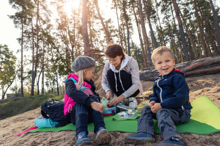 Young adult caucasian beautiful mother enjoy having fun eating snack on picnic with two cute little children in pine forest outdoors on warm autumn day. Family nature outdoor recreation lifestyleの写真素材