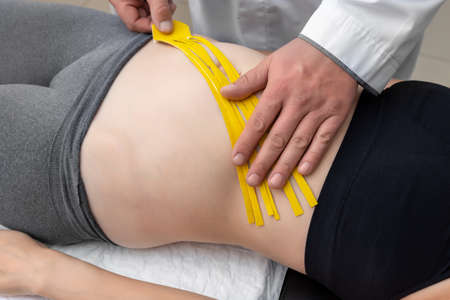 Male physiotherapist applying yellow adhesive elastic therapeutic tape on female abdominal muscle of young adult slim sporty woman lying on couch doctor office. Injury rehabilitation and healthcareの写真素材