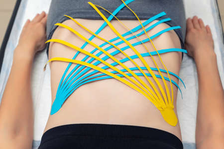 Flat lay above view applying blue adhesive elastic therapeutic tape on female abdominal muscle of young adult slim sporty woman lying on couch doctor office. Injury rehabilitation and healthcareの写真素材