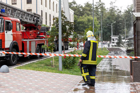 Firemen un uniform standing behind fire tape and many fire engine trucks with ladder at accident in...