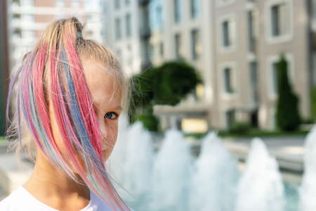 Cute adorable caucasian little kid girl with multicolored pastel chalk pained dyed blond hair on city street with fountain background. Female child portrait pink colorful hairstyle smiling outdoorsの写真素材