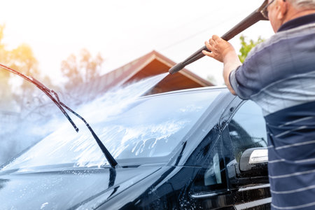 Senior male person wash car van with high pressure water equipment pump at home backyard outdoors on bright shiny summer day. Vehicle covered with foam chemical detergents during carwash self serviceの写真素材