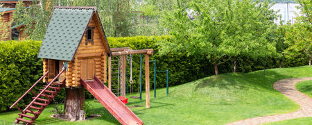 Small wood log playhouse hut with stairs ladder and wooden slide on children playground at park or house yard. Green grass lawn garden and paved pathway background on bright sunny day. Panoramic viewの写真素材