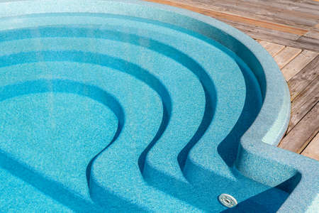 New modern fiberglass plastic swimming pool entrance step with clean fresh refreshing blue water on bright hot summer day at yard or resort hotel spa area. Wooden flooring deck of teak or larch boardの写真素材