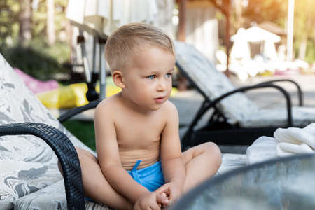 Cute adorable caucasian blond little pensive kid boy enjoy having fun relaxing sitting on sunbed in yard garden near pool sea beach. Child chilling outdoor on sunny day. Toddler sunbathing outsideの写真素材