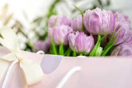 Close-up detail of fresh beautiful present of tender violet peony tulips bouquet and paper gift bag with romantic bow. Wedding or anniversary celebration surprise background. Pastel floral decorationの写真素材