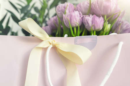 Close-up detail of fresh beautiful present of tender violet peony tulips bouquet and paper gift bag with romantic bow. Wedding or anniversary celebration surprise background. Pastel floral decorationの写真素材