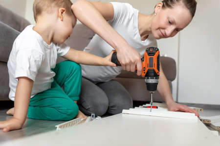 Cute adorable caucasian toddler boy kid sit on floor and help mom assembling furniture shelf with power screwdriver tool. Young adult mum with funny little child enjoy playing at home indoorsの写真素材