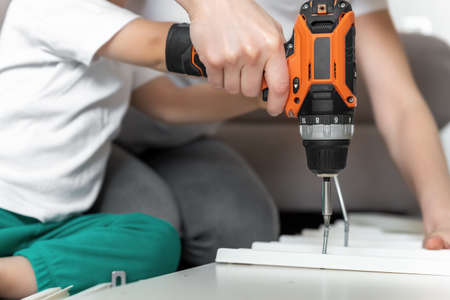 Close-up detail toddler boy kid sit on floor and help mom assembling furniture shelf with power...