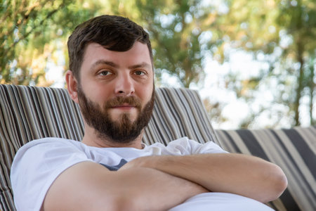 Close-up portrait of young adult cuacasian bearded man enjoy chilling on sofa at yard outdoors on sunny warm summer day. Happy smiling male person rest in park outsideの写真素材