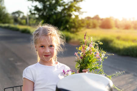 Close-up portrait of little cute caucasian blond smiling kid girl holding bouquet of salvia sage wild flower sitting in bicycle rack grass meadow country road summer day. Nature summertime activitiesの写真素材