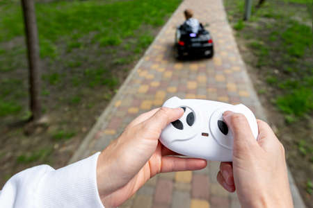 Parent hand holding remote controller of electric powered battery car with little kid boy enjoy having fun riding toy asphalt path road city park at summer day. Mother child safety and care conceptの写真素材