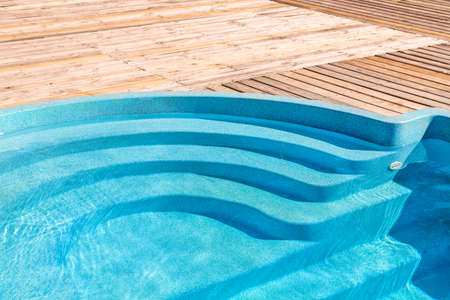 New modern fiberglass plastic swimming pool entrance step with clean fresh refreshing blue water on bright hot summer day at yard or resort hotel spa area. Wooden flooring deck of teak or larch boardの写真素材