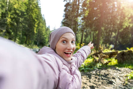 Young adult caucasian beautiful happy smiling woman making selfie shot by cell phone pointing finger backward during walk in scenic green woods pine forest river. Person enjoy walking nature outdoorsの写真素材