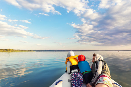 Bacl view two cute adorable little caucasian sibling together with mother wearing lifejacket vest having fun enjoy riding inflatable motorboat river against scenic nature landscape. summer recreationの写真素材
