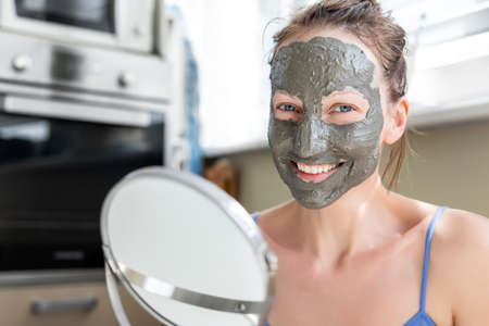 Young adult happy beautiful caucasian woman applying natural gray clay face mask at home against window and looking in mirror. Female person making self facial cleansing moisturizing treatment careの写真素材