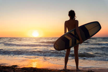 Back view young adult slim sporty female surfer girl with surfboard silhouette stand at ocean coast wave against warm sunrise or sunset sun. Sport healthy carefree freedom lifestyle vacation conceptの写真素材