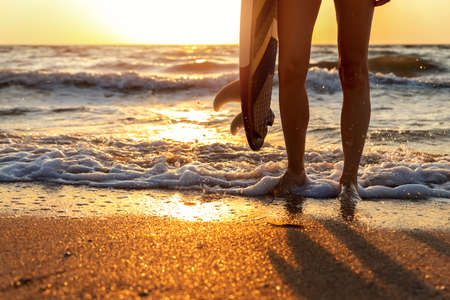 Close-up bottom pov view young adult female surfer girl legs with surfboard stand at ocean coast wave against warm sunrise or sunset sun. Sport healthy carefree freedom lifestyle vacation conceptの写真素材