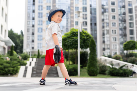 Cute adorable caucasian 3-4 years blond little kid boy in stylish shorts shirt and hat hold soft teddy toy enjoy playing outdoors at urban city center downtown area. Funny child portraitの写真素材