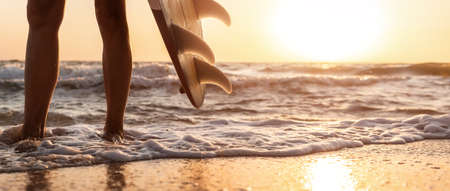 Close-up bottom pov view young adult female surfer girl legs with surfboard stand at ocean coast wave against warm sunrise or sunset sun. Sport healthy carefree freedom lifestyle vacation concept.の写真素材