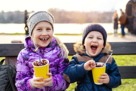 Two cute adorable siblings children sitting on bench drink delicious yummy hot chocolate, tea cocoa from paper cups during walk at city street park or backyard outdoors. Brother and sister enjoy funの写真素材