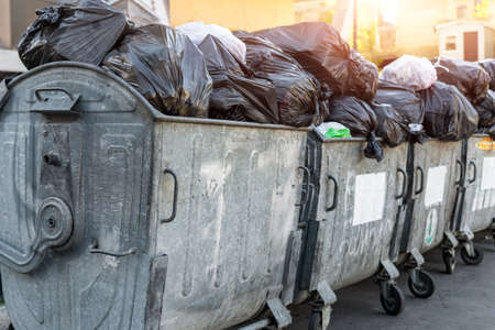 Rows of many big metal overloaded dumpster cans full of black plastic trash litter bags near residential building at city downtown or suburban area. Non-recyclable sorting garbage collectingの写真素材
