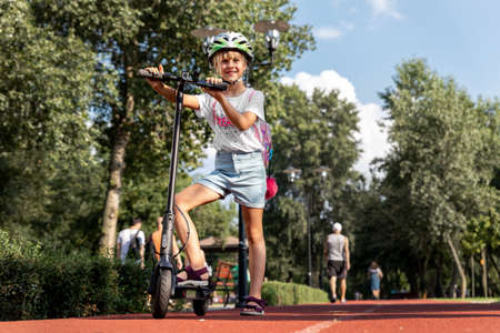 Profile view portrait of cute blond little caucasian school girl wear helmet enjoy having fun riding electric scooter city street park outdoors on sunny day. Healthy sport children activities outsideの写真素材