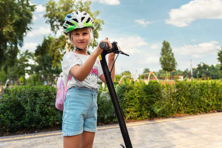 Profile view portrait of cute blond little caucasian school girl wear helmet enjoy having fun riding electric scooter city street park outdoors on sunny day. Healthy sport children activities outsideの写真素材